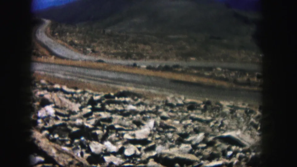Grainy 1958 Colorado 8mm view of stones, roadside dirt, and a curving road through rugged terrain.