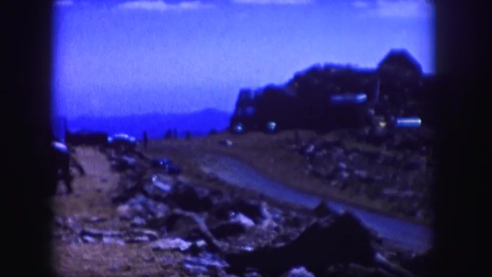 Blue-tinted 1958 Colorado 8mm frame showing a mountain road, rocky shoulder, distant hills, and tiny people near the road.