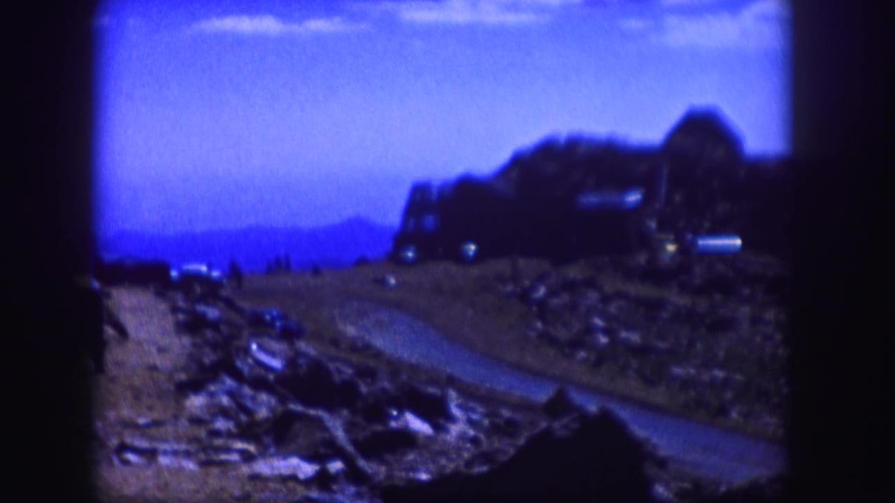 Blue-tinted mountain road curves through rocky terrain toward a distant hillside structure.