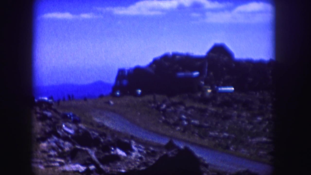 Blue-cast 1958 Colorado home-movie frame with a winding road, rocky slope, distant structure, and tiny visitors near the horizon.