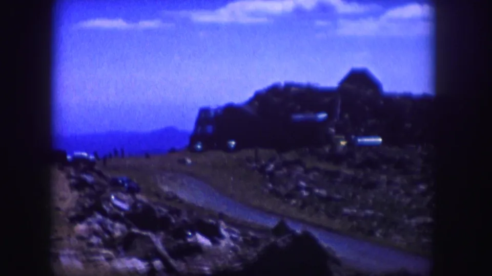 1958 Colorado 8mm still with strong blue tint, soft focus, rocky roadside terrain, a curving road, and distant hillside structure.