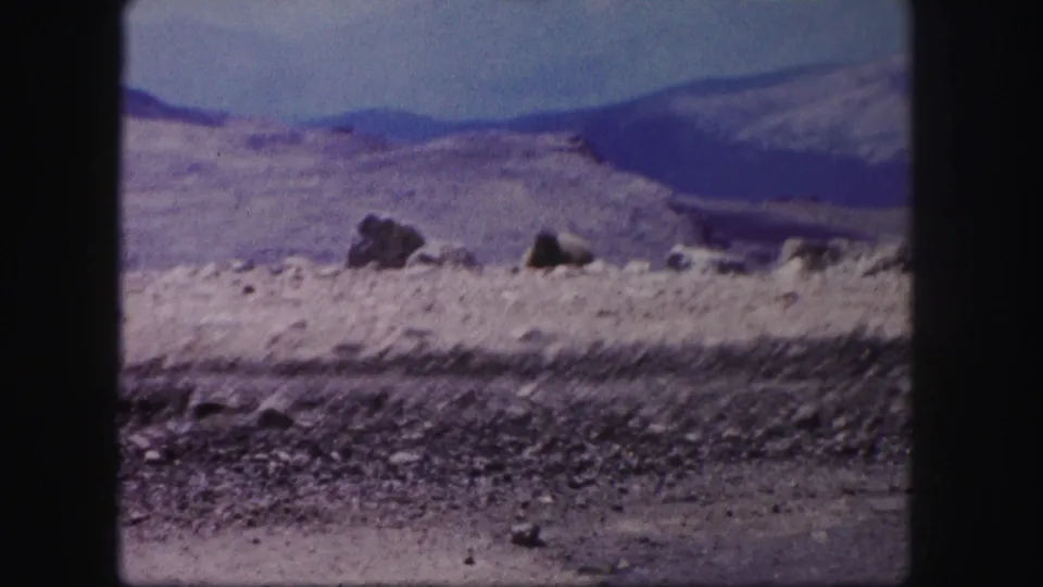 1958 Colorado 8mm frame showing a rocky roadside foreground, pale desert rise, scattered boulders, and blue mountain ridges.