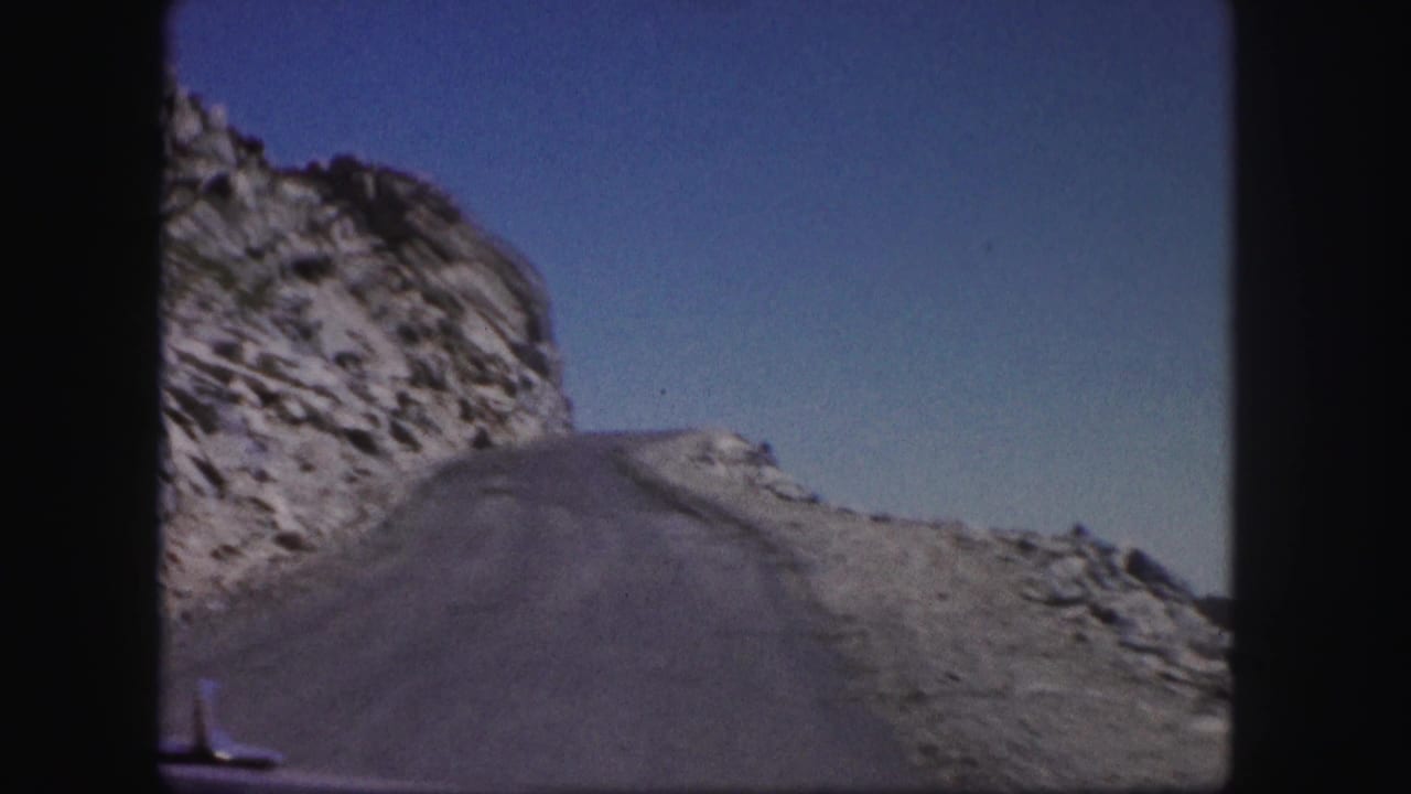 Mountain road curves uphill beside a rocky slope under a clear blue sky.