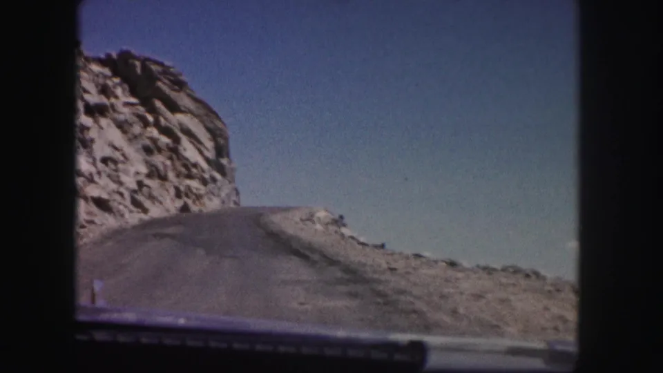 1958 Colorado home-movie frame showing a paved road bending past a rocky outcrop from a moving vehicle.
