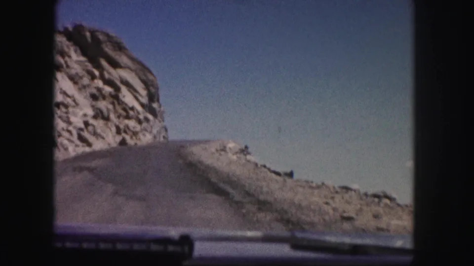 Colorado mountain-driving POV on 8mm film, with the road, rocky slope, and car hood visible in frame.