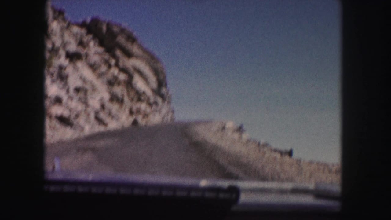 1958 Colorado 8mm driving view from inside a car, with a purple hood, road edge, rocky hillside, and blue sky.