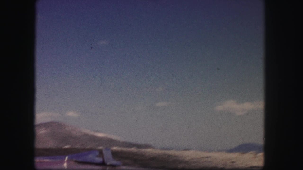 1958 Colorado mountain drive view with dry slopes, blue sky, and a partial vehicle edge in soft 8mm color.