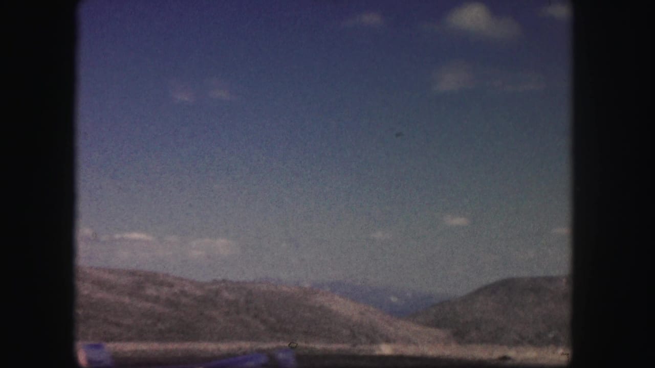 Colorado mountain terrain appears beyond the road in a soft, grainy 8mm frame from a moving vehicle.