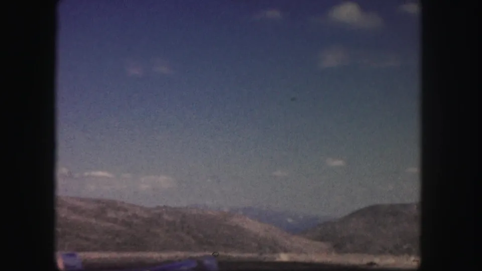 Colorado mountain terrain appears beyond the road in a soft, grainy 8mm frame from a moving vehicle.