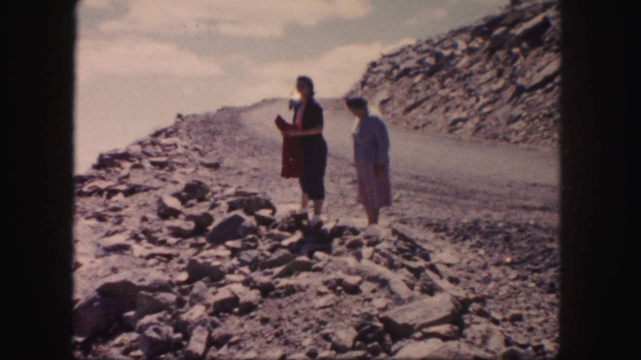 Two adult women in 1950s-style clothing stand on a rocky Colorado roadside, facing the open valley and distant terrain.