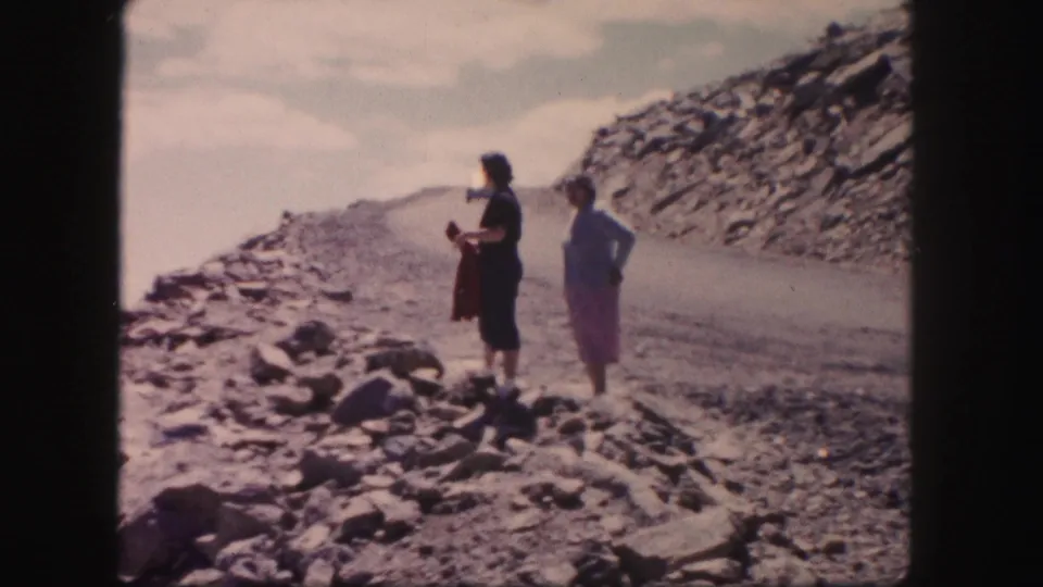 The Colorado home-movie frame shows two adult women on an unpaved rocky road, with arid terrain stretching away beside them.