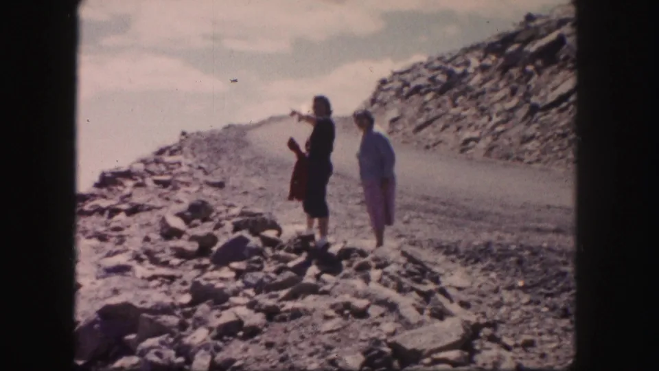 On a rocky Colorado roadside, one adult woman gestures toward the distant valley while another stands nearby in the 1958 home-movie footage.