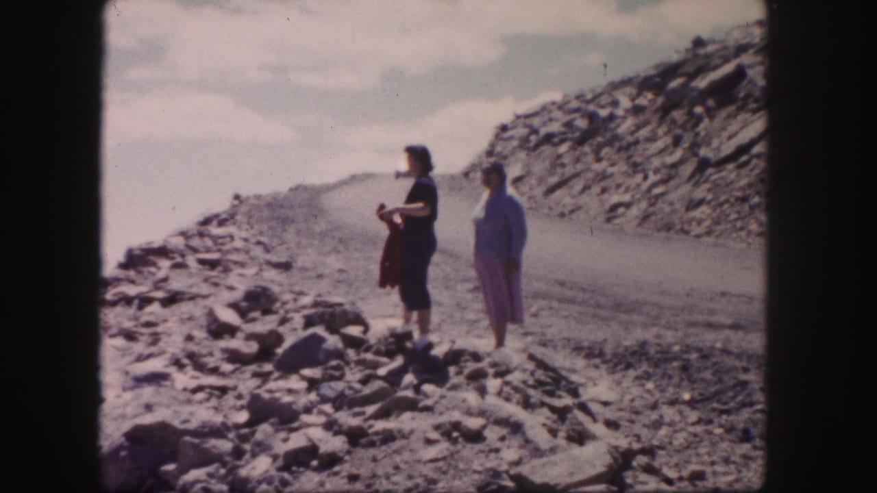 Two women stand on a rocky roadside with a winding mountain road behind them.