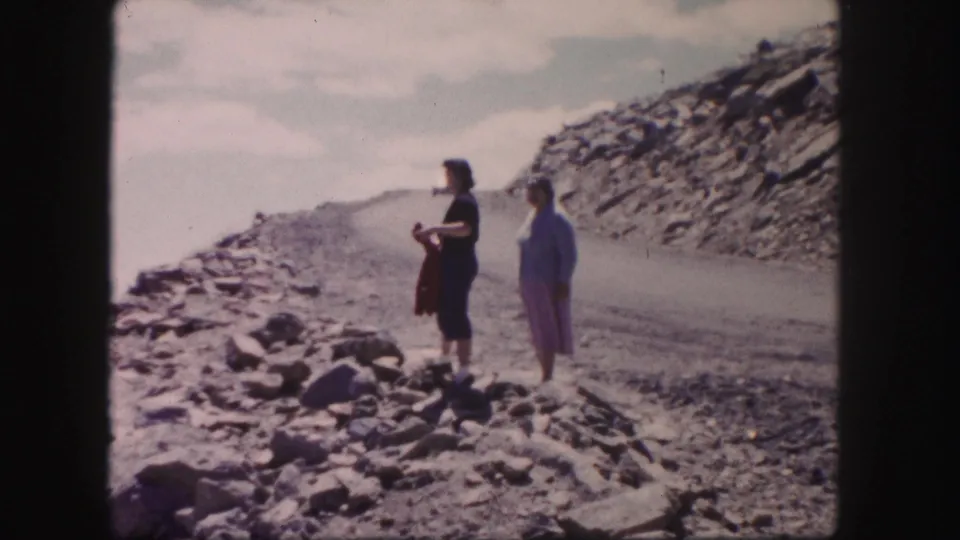 Two women stand apart on a rough Colorado mountain road, with loose rock and a sloped hillside behind them.