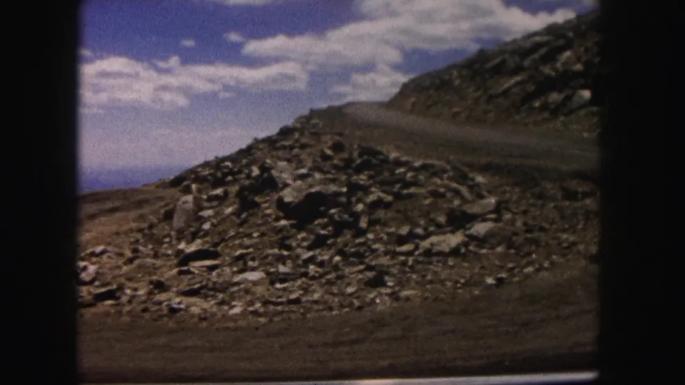 A wider Colorado mountain-road view shows loose rocks, a curving dirt track, distant terrain, and 8mm frame edges.