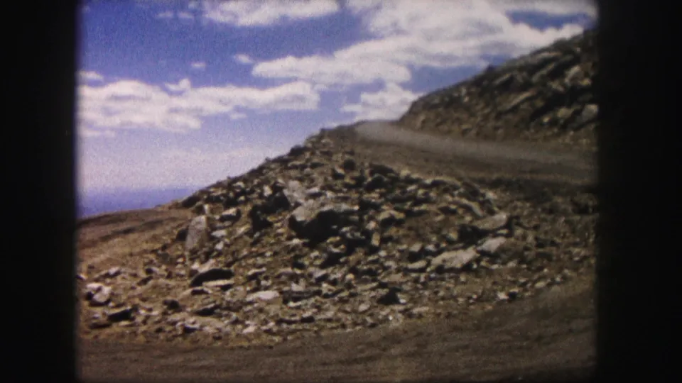 A sunlit Colorado mountain road curves around a rubble-strewn slope, with distant landscape visible beyond the rocks.
