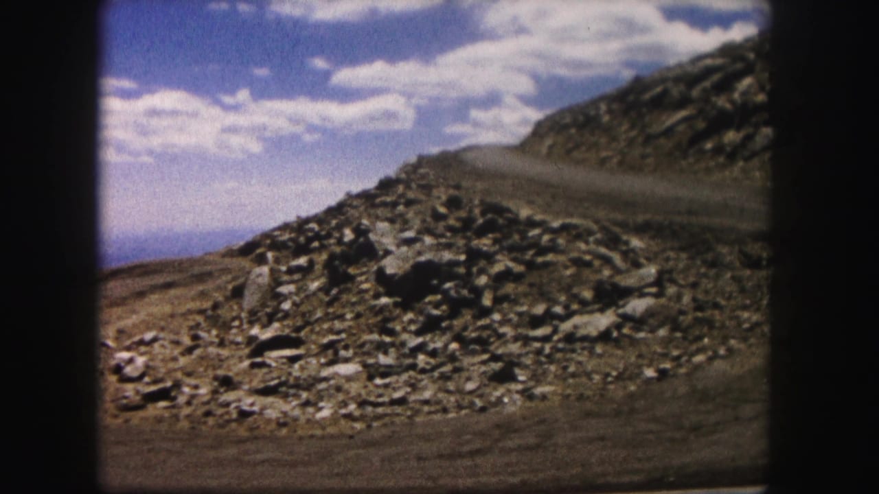 1958 Colorado 8mm frame showing a rocky slope, dry roadside ground and blue cloudy sky within dark film borders.