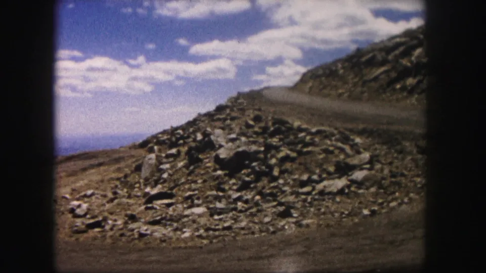 1958 Colorado travel footage frame with a rocky hillside, unpaved road edge, distant horizon and scattered white clouds.