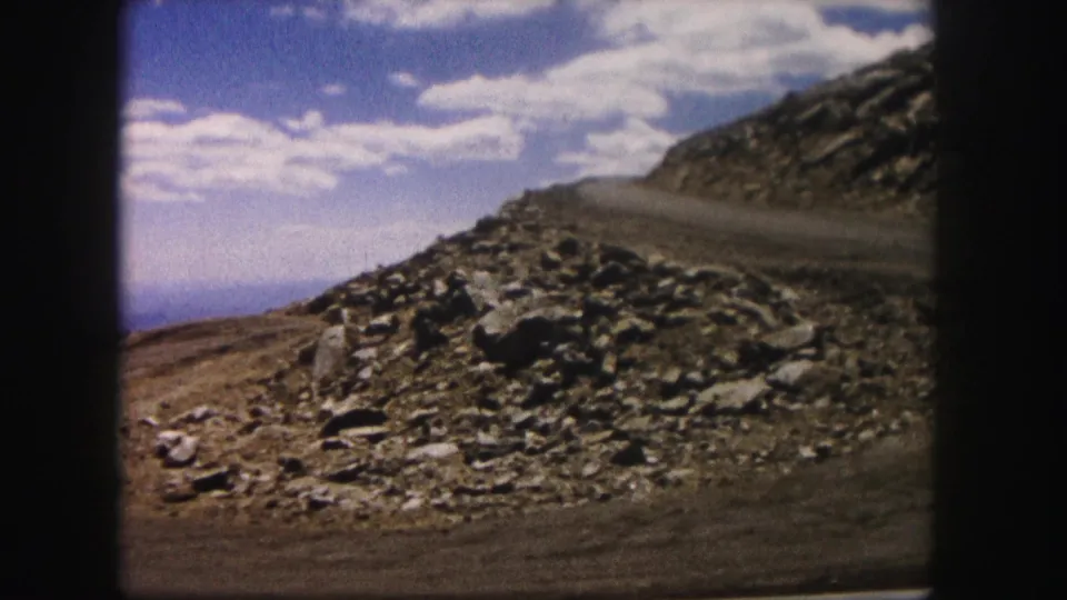 1958 Colorado 8mm frame showing dry roadside terrain, loose rocks, a sloping hill and a bright sky with scattered clouds.