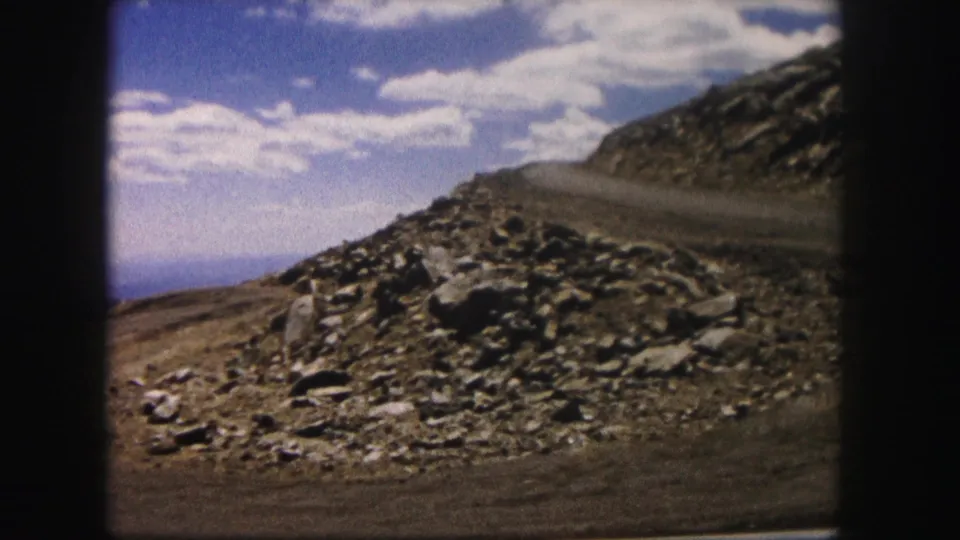 1958 Colorado 8mm frame of a rocky mountain roadside, dirt track, dry slope and clouded blue sky framed by dark film edges.
