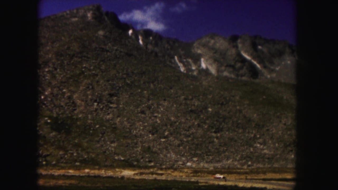 1958 Colorado 8mm frame showing a rocky mountain slope, snow patches, and a dirt road at the base under a blue sky.