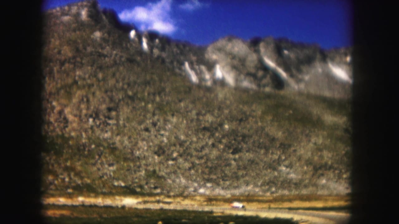 Mountain range with snow patches and a small white vehicle on a distant road.