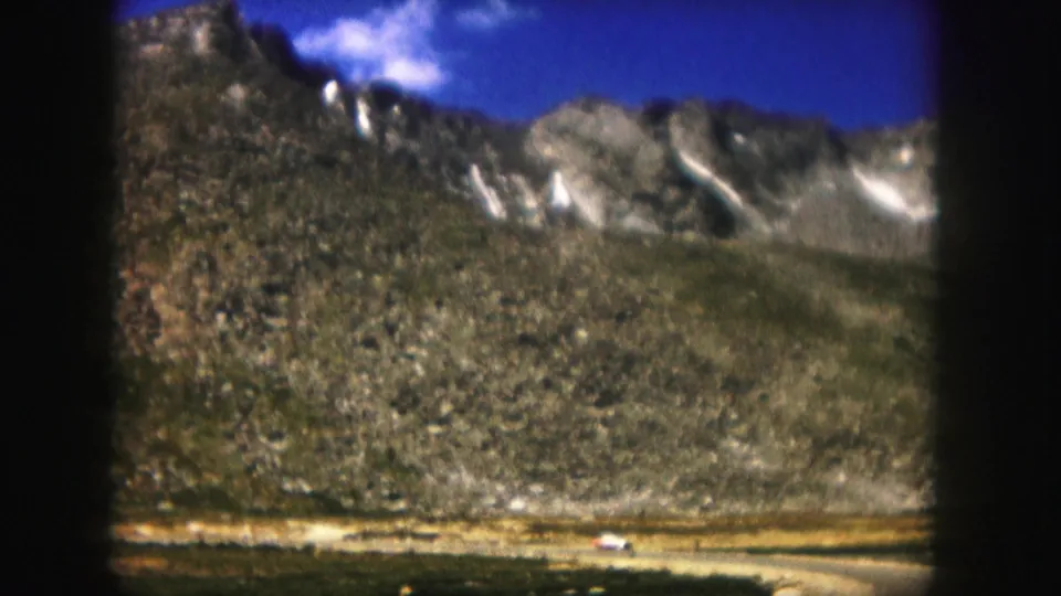 1958 Colorado home-movie frame showing rocky mountain slopes, pale snow streaks, and open terrain near a distant road.