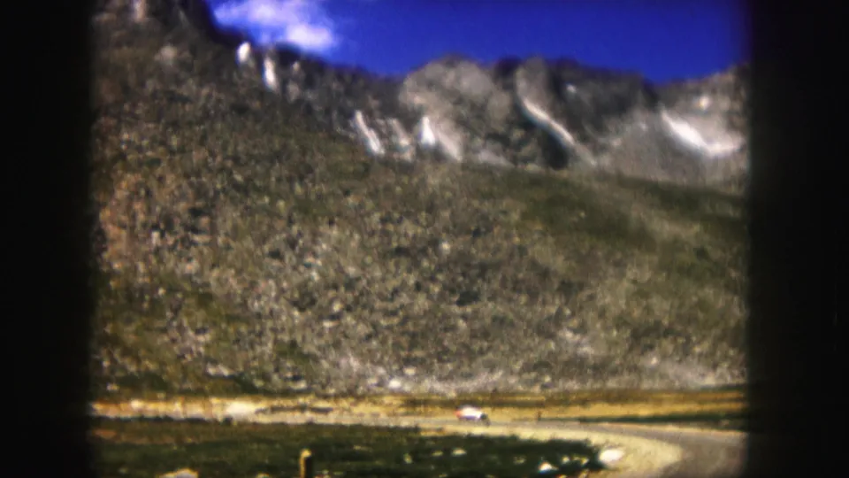 1958 Colorado 8mm frame showing a winding dirt road below rocky mountains, with a small white vehicle in the distance.