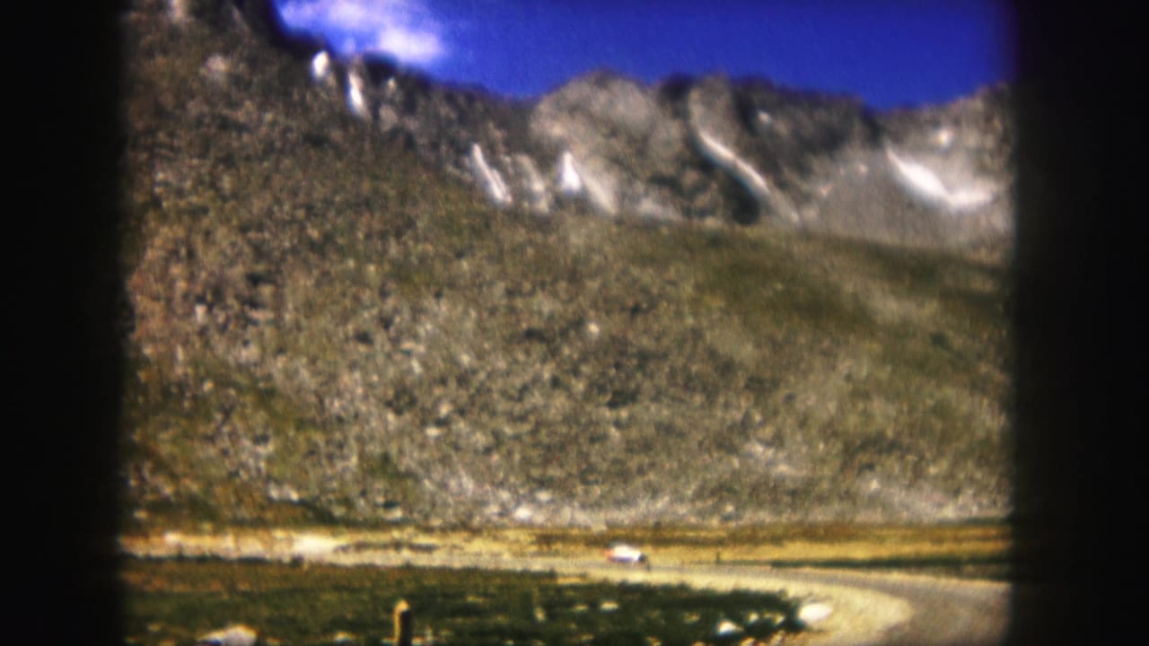 1958 Colorado 8mm frame showing a small white car on a winding road beneath rocky mountain slopes and patches of snow.