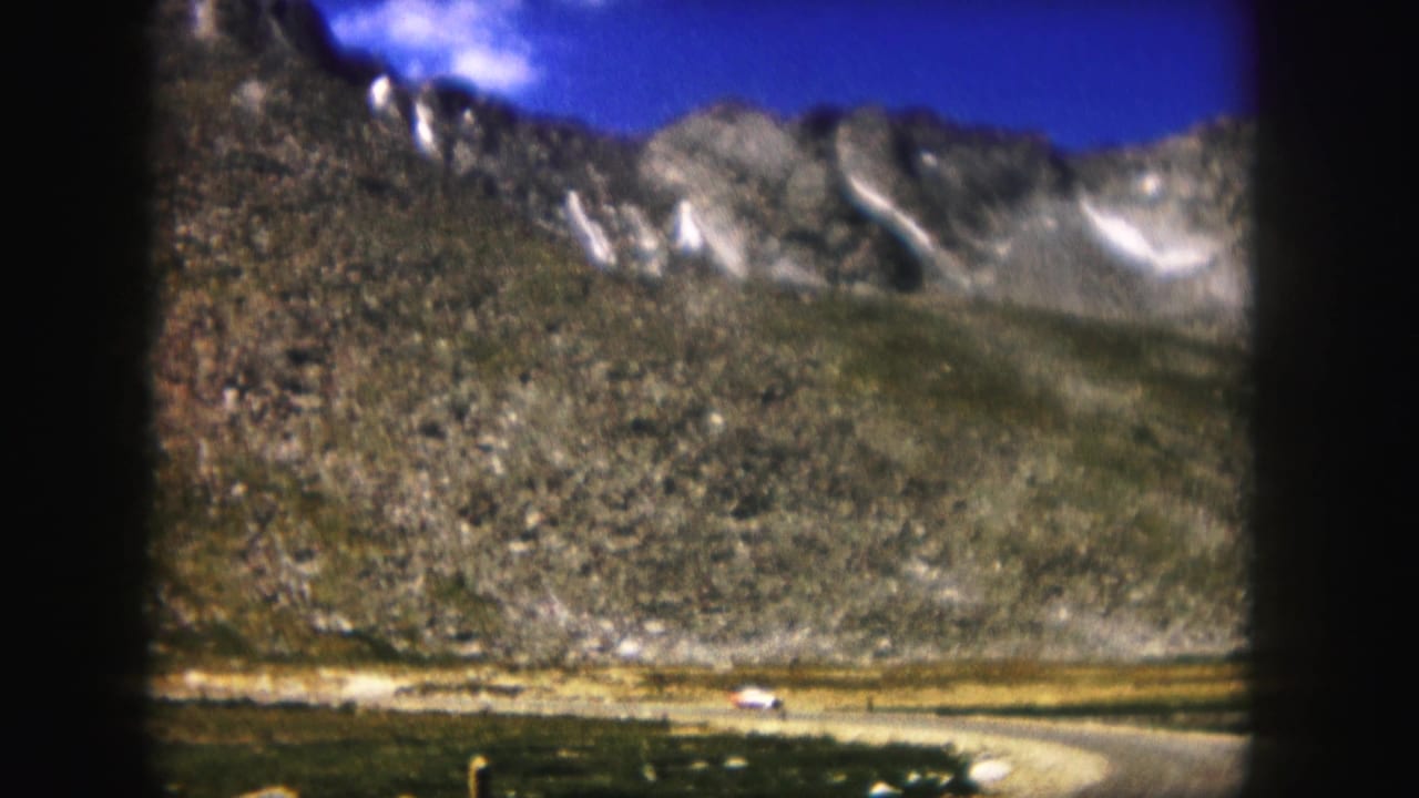 Curving road and small white car at the base of a rocky mountain slope.