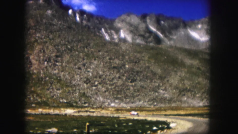 1958 Colorado frame showing a small white car near the bend of a road below rugged mountain slopes.