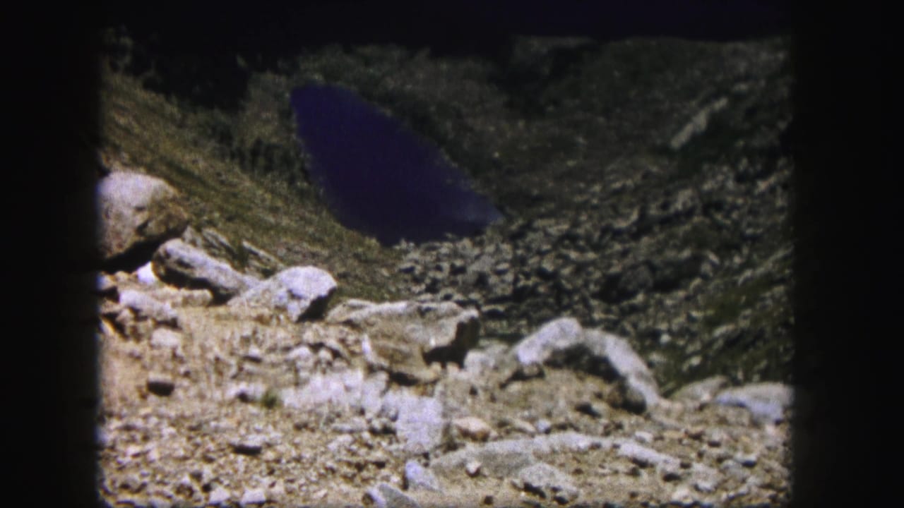 Rock-strewn mountain slope leading toward a dark alpine lake or pond.