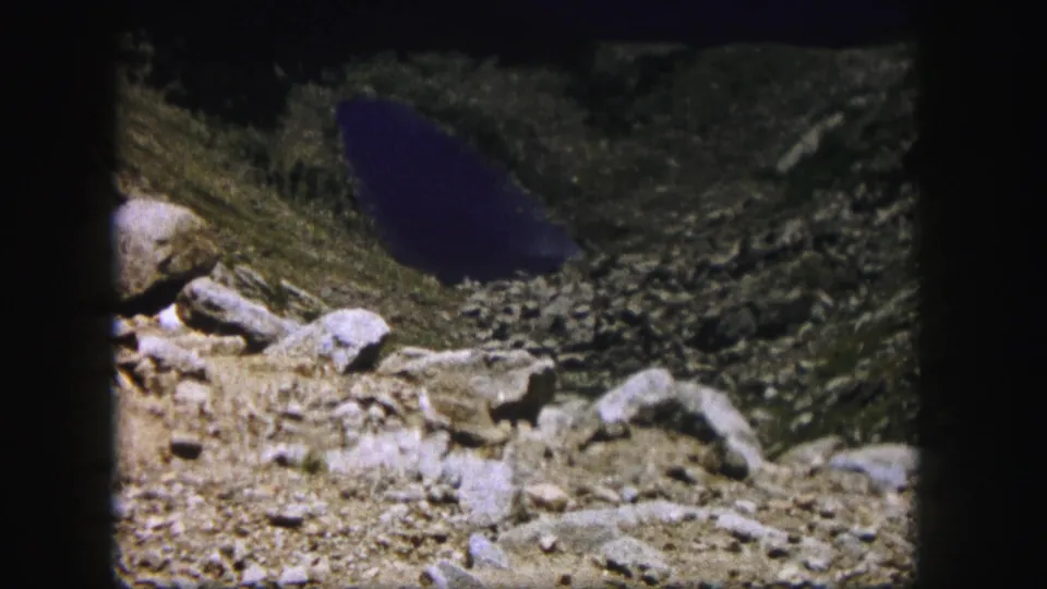 1958 Colorado 8mm frame showing boulders, gravelly ground, sparse vegetation, and dark alpine water in a mountain basin.