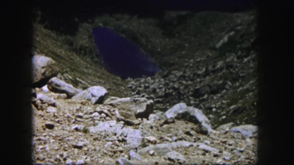 1958 Colorado 8mm frame of a rocky mountain basin, with pale boulders, loose debris, and dark water in the background.