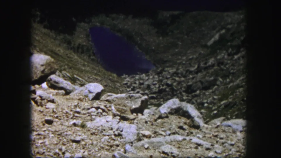 1958 Colorado 8mm frame showing loose rock, large boulders, sparse vegetation, and dark alpine water in rugged terrain.