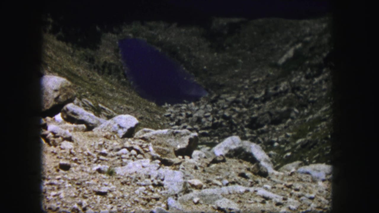 Distant blue lake below a rocky mountain ledge with stones in the foreground.