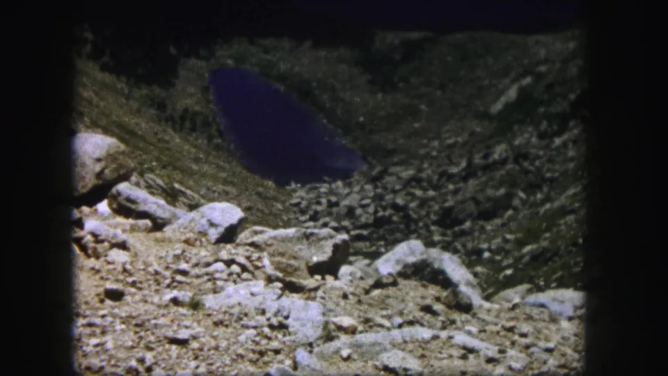 1958 Colorado 8mm frame showing a dark blue mountain lake below a rocky ledge and sloped basin.