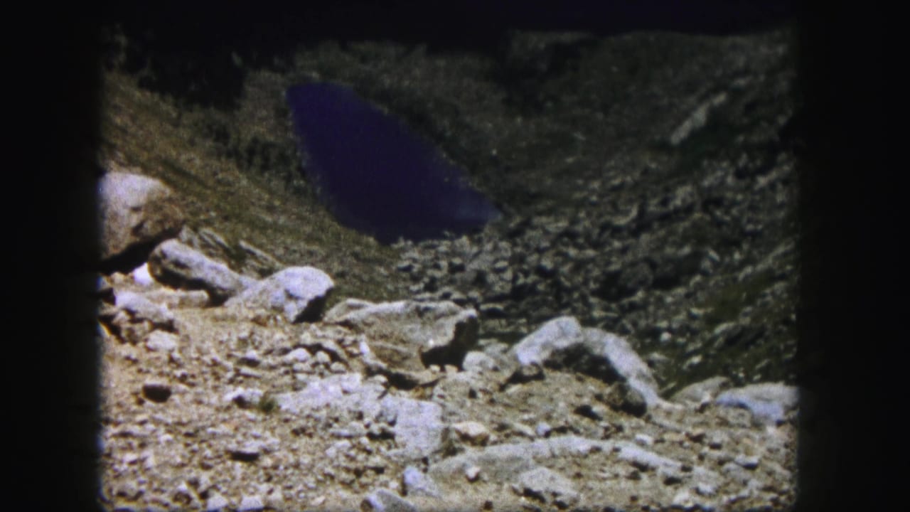 A Colorado mountain lake appears below the rocky overlook, with 8mm grain and dark frame edges visible.