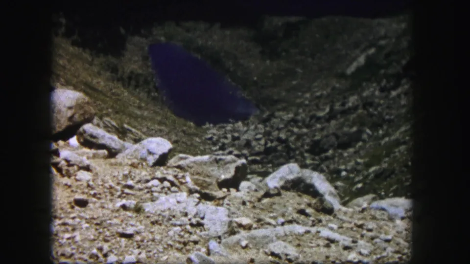 A Colorado mountain lake appears below the rocky overlook, with 8mm grain and dark frame edges visible.