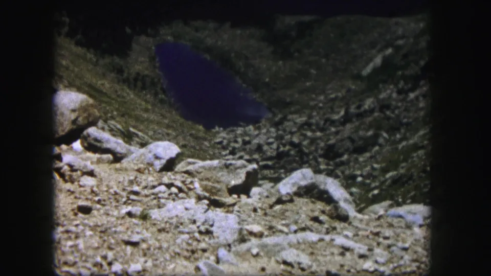 1958 home-movie view of a dark blue lake beyond foreground rocks on a Colorado mountain slope.