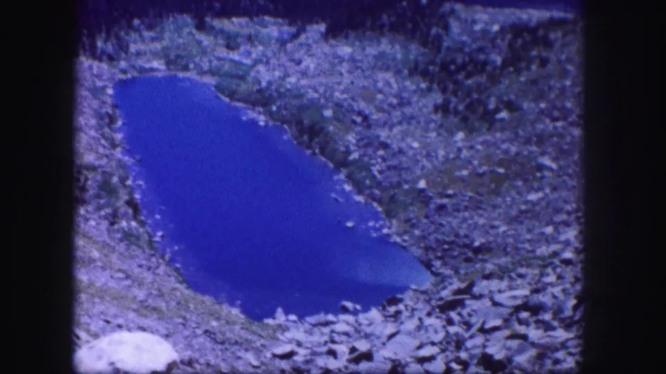 Zoomed-in 1958 Colorado 8mm frame with a bright blue lake ringed by gray and brown rocks.