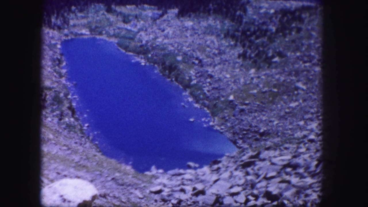 1958 Colorado 8mm frame showing a vivid blue alpine pond surrounded by gray rocks and sparse vegetation.