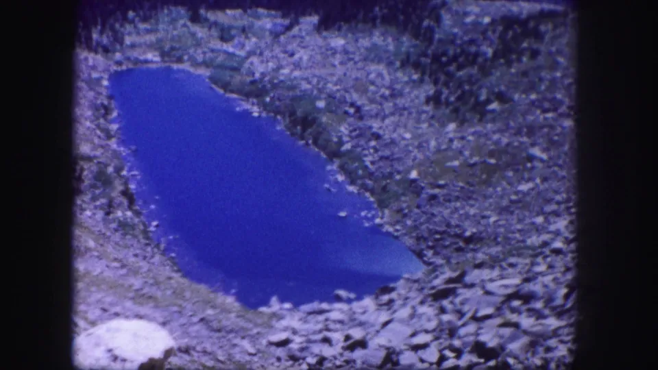 1958 Colorado 8mm frame showing a vivid blue alpine pond surrounded by gray rocks and sparse vegetation.