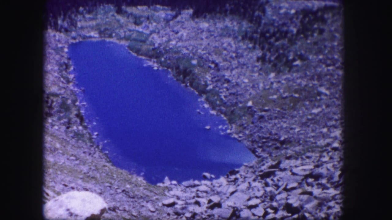 1958 Colorado 8mm frame with a static view of a deep blue pond edged by rocky terrain.