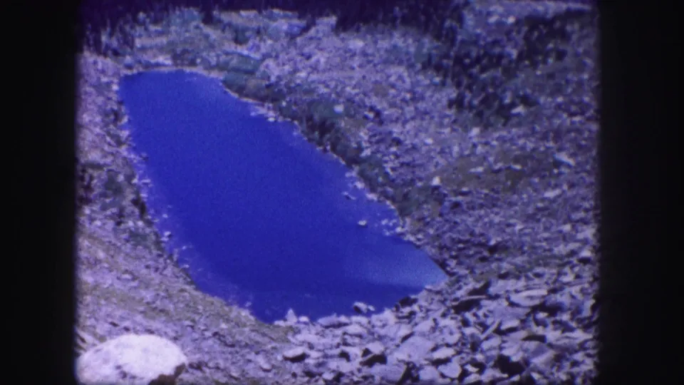 1958 Colorado 8mm frame with a static view of a deep blue pond edged by rocky terrain.
