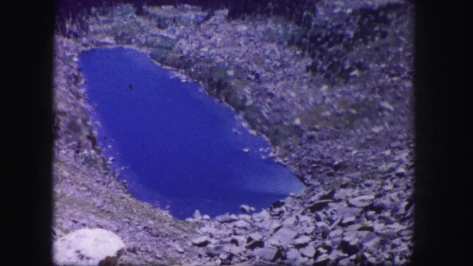 1958 Colorado 8mm frame showing the same alpine pond from a high, slightly shifted home-movie viewpoint.