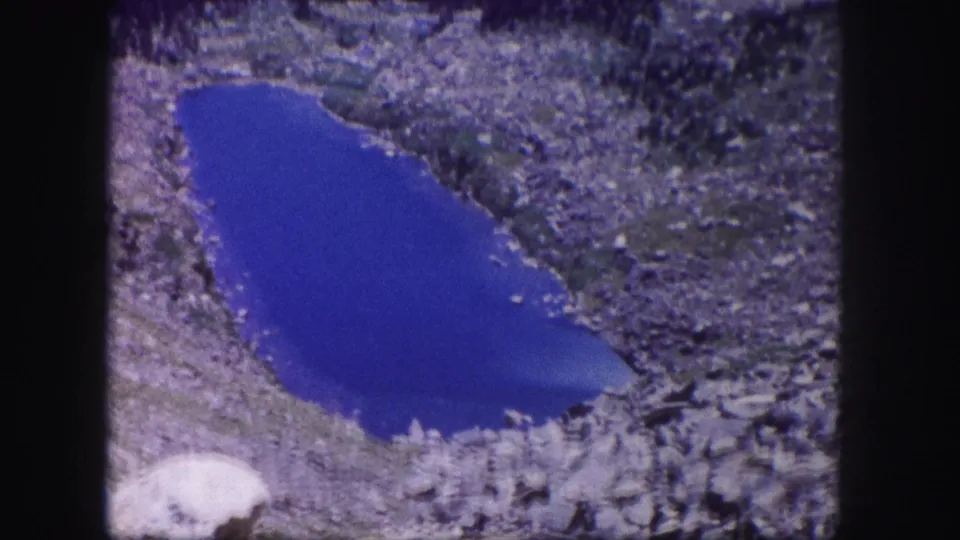 1958 Colorado 8mm frame of a vivid blue mountain pond, rocky shoreline, and soft film texture.