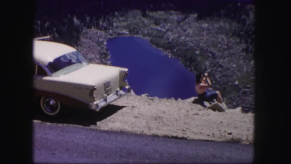 1958 Colorado 8mm frame of a two-tone 1950s sedan stopped near an alpine lake while a woman sits on the rocks with a camera.