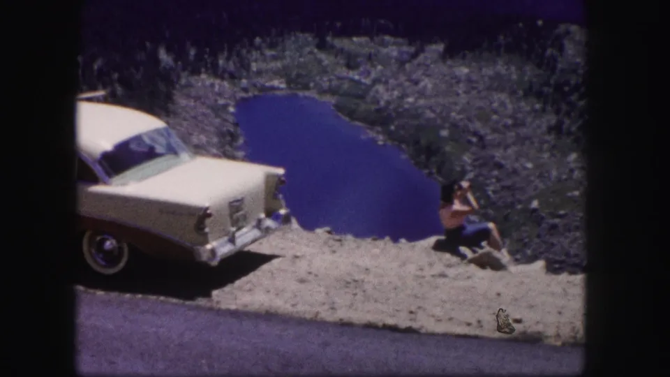 Colorado 1958 home-movie still showing a classic white sedan, rocky lakeside shoulder, and a woman seated with a camera.