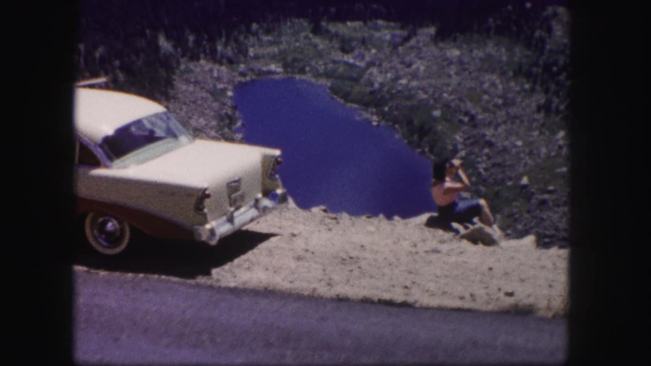 In 1958 Colorado footage, a woman sits by a rocky overlook above a blue lake while a cream and maroon sedan is parked nearby.